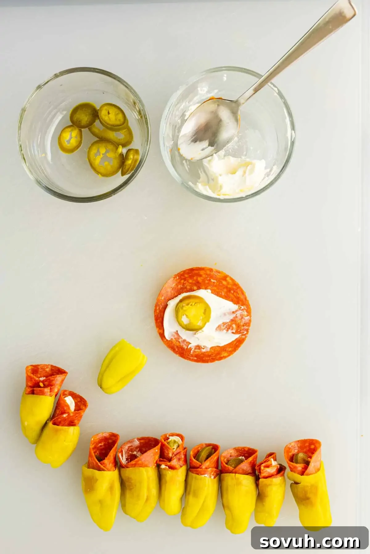 Air Fryer Stuffed Pepperoncinis with Pepperoni and Hot Honey 5 Two glass bowls with sliced jalapeños and cream cheese, a slice of pepperoni with cream cheese and a jalapeño on top, and several assembled pepperoni stuffed pepperoncinis snacks on a white surface.