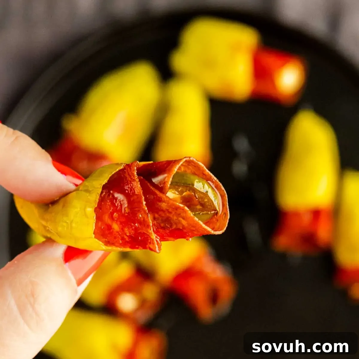 Air Fryer Stuffed Pepperoncinis with Pepperoni and Hot Honey 6 A hand holds a yellow pepperoni stuffed pepperoncini filled with sliced meat, with more air fryer stuffed pepperoncinis arranged on a black plate in the background.