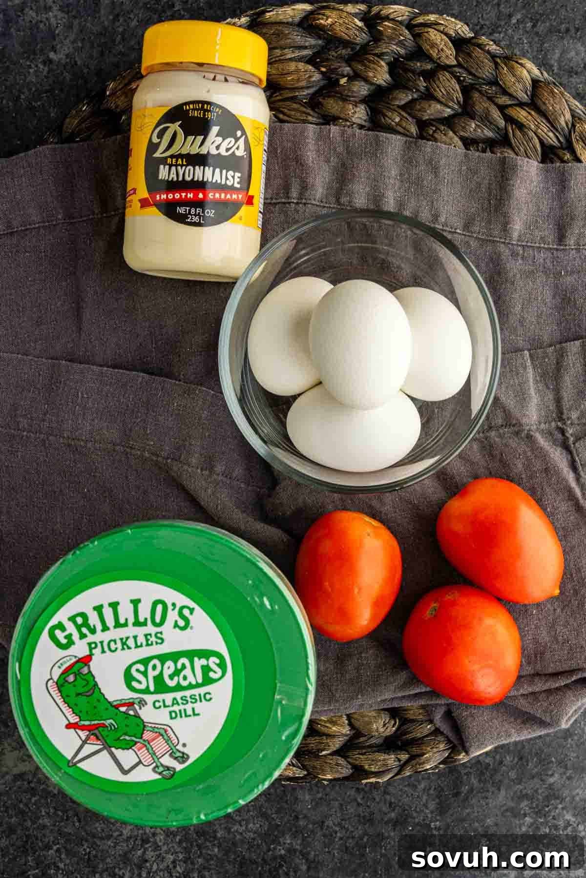Viral TikTok Layered Egg Salad Dip Recipe 3 A jar of mayonnaise, a bowl of four eggs, three Roma tomatoes, and a container of dill pickles arranged on a dark cloth and woven mat—perfect ingredients for a delicious layered egg salad dip.