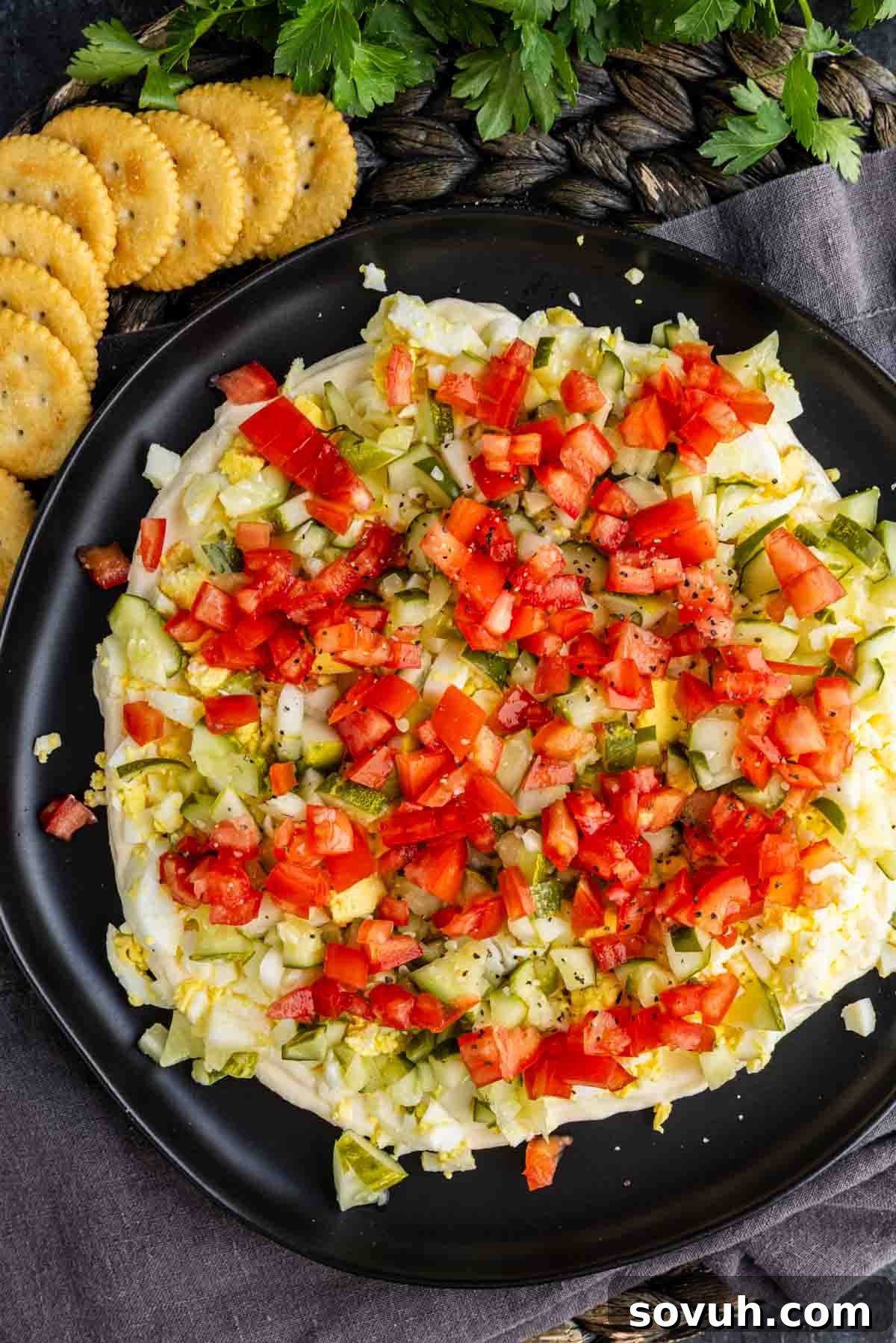 Viral TikTok Layered Egg Salad Dip Recipe 5 A black plate holds layered egg salad dip covered with chopped tomatoes, pickles, and onions, surrounded by round crackers. Parsley is partially visible at the top of the image.