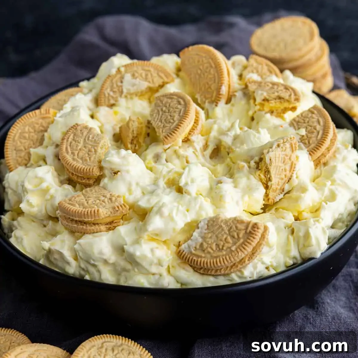 Lemon Cookie Salad: Bright Midwestern Dessert Twist 2 A black bowl filled with a creamy lemon fluff dessert mixture, topped with pieces of golden sandwich cookies, with more cookies scattered around the bowl.