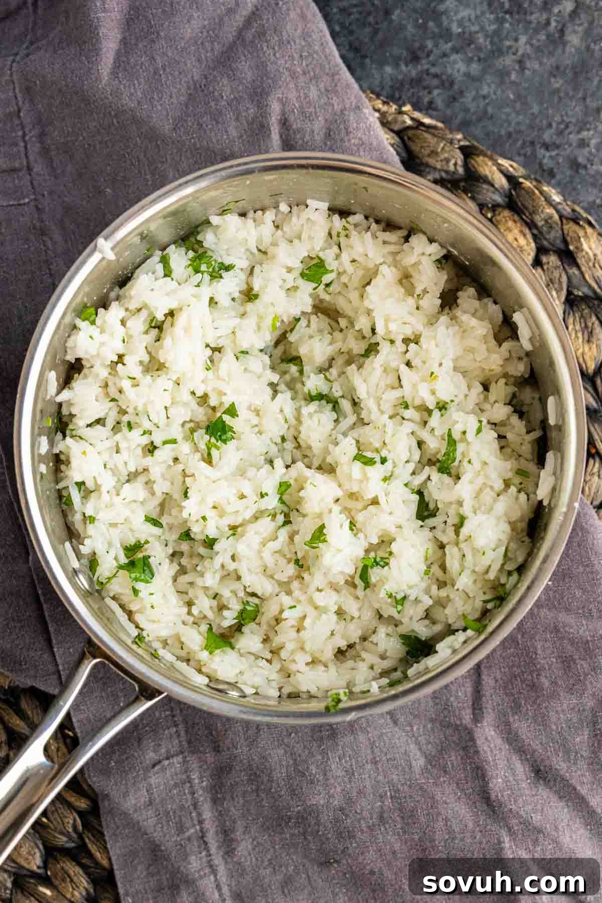Fluffy Coconut Lime Rice Recipe with Fresh Citrus Flavor 4 A stainless steel pot filled with easy coconut lime rice, cooked white rice mixed with chopped herbs, placed on a gray cloth and wicker mat.