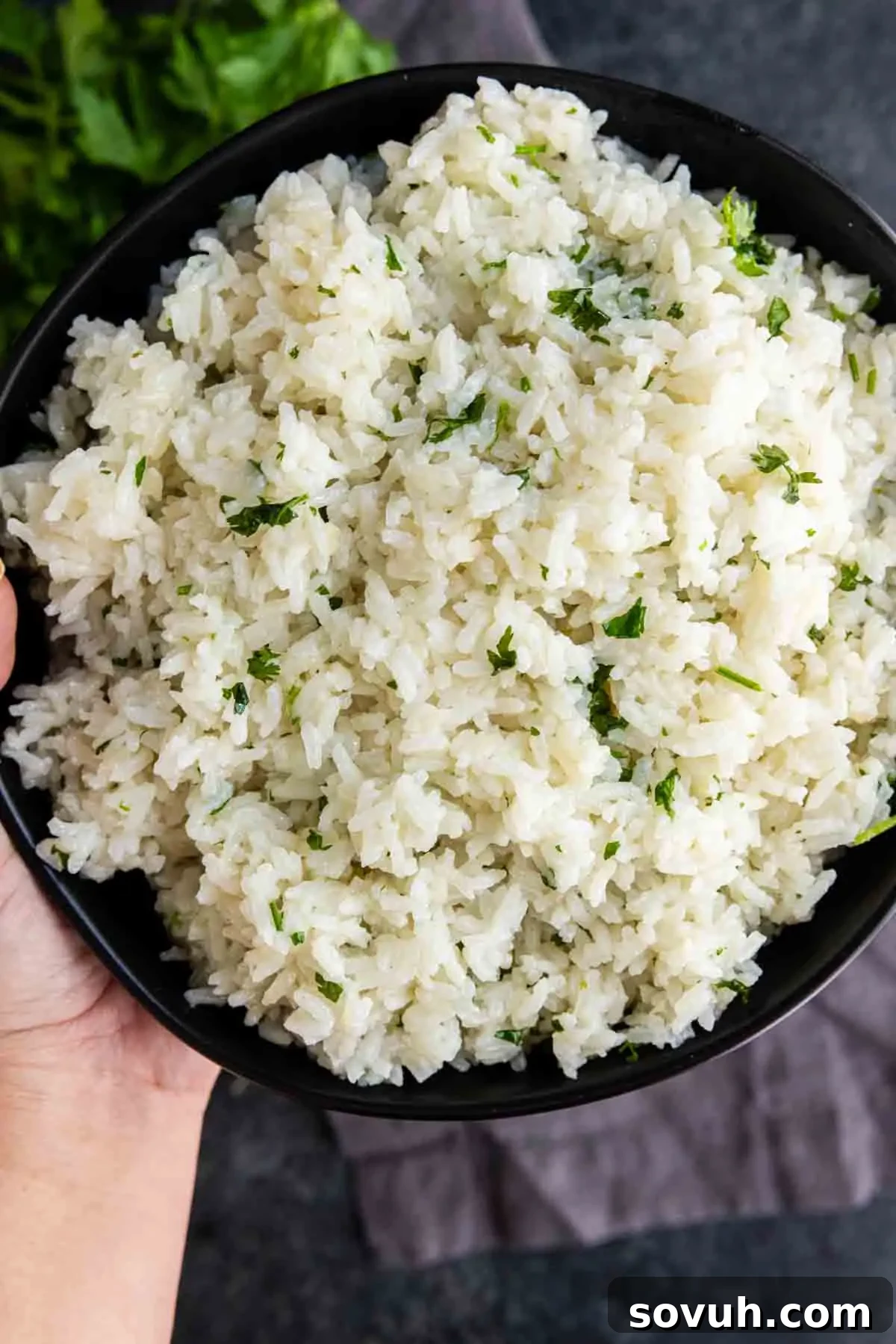 Fluffy Coconut Lime Rice Recipe with Fresh Citrus Flavor 6 A bowl of easy coconut lime rice garnished with chopped herbs, held in one hand over a dark surface.