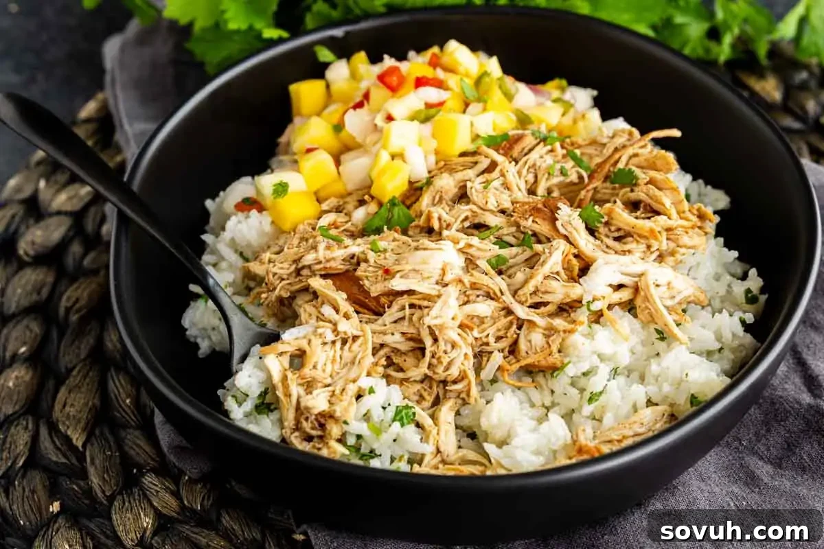 Slow Cooker Jerk Chicken Recipe 2 A black bowl filled with shredded chicken over white rice, topped with diced mango salsa, with a black spoon on the side.