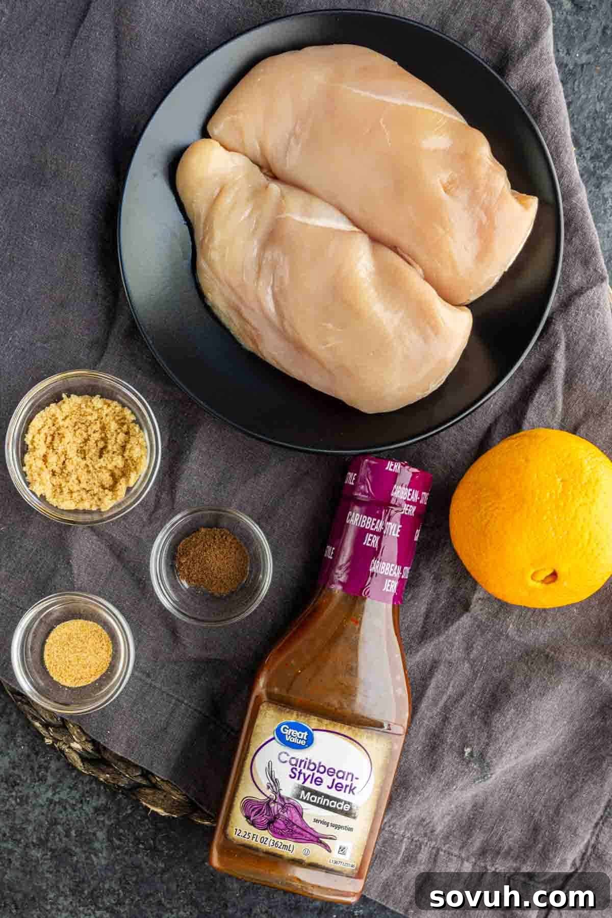 Slow Cooker Jerk Chicken Recipe 3 Two raw chicken breasts on a black plate, a bottle of Caribbean-style jerk marinade, an orange, brown sugar, allspice, and garlic powder arranged on a gray cloth.