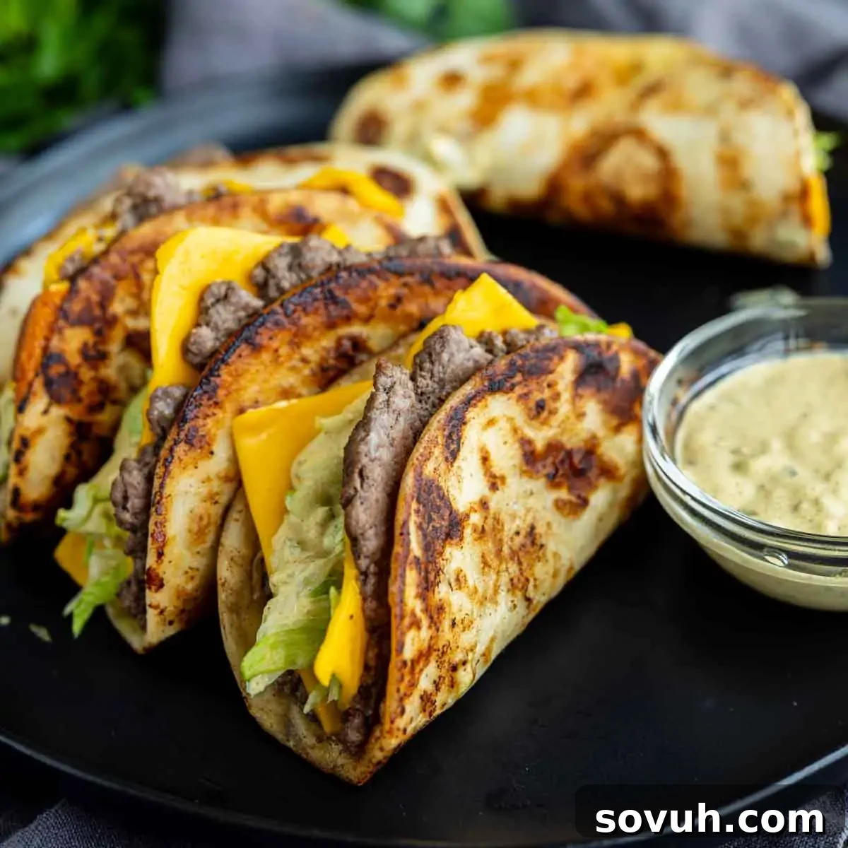 Big Mac-Style Smash Tacos with Homemade Big Mac Sauce 2 Three grilled tacos filled with beef, lettuce, and cheddar cheese are arranged on a black plate next to a small bowl of dipping sauce—an easy weeknight dinner inspired by the viral taco recipe trend.