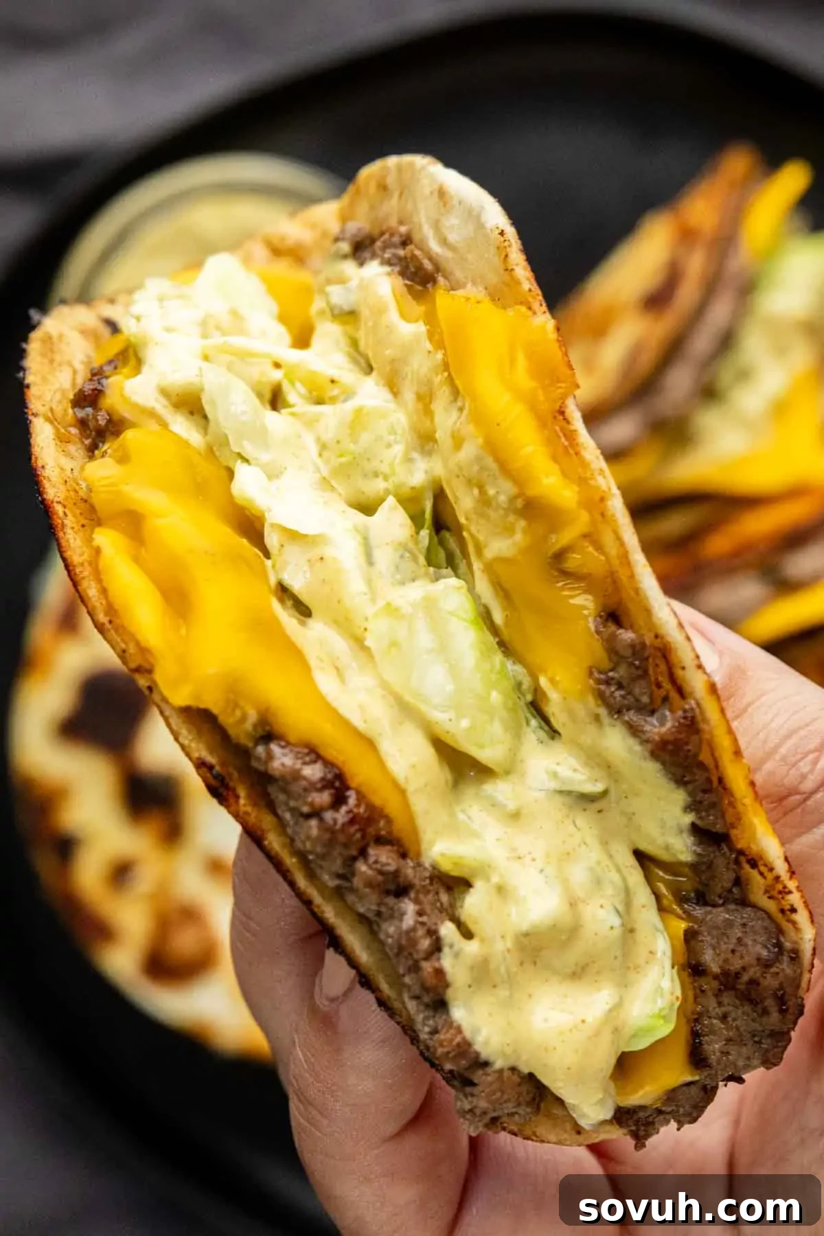 Big Mac-Style Smash Tacos with Homemade Big Mac Sauce 5 A hand holds a folded flatbread filled with beef, cheddar cheese, lettuce, and a creamy sauce—an irresistible take on the viral taco recipe—with more sandwiches on a black plate in the background.