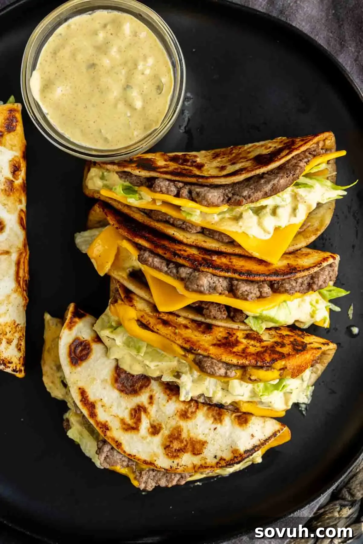 Big Mac-Style Smash Tacos with Homemade Big Mac Sauce 6 Three folded tortillas filled with ground beef, cheddar cheese, lettuce, and sauce are arranged on a black plate next to a small bowl of creamy dipping sauce—an easy weeknight dinner inspired by the viral taco recipe trend.