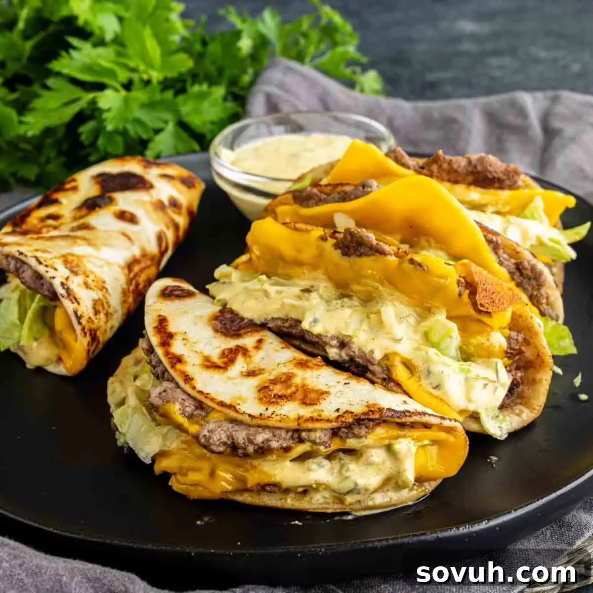 Big Mac-Style Smash Tacos with Homemade Big Mac Sauce 7 Three cheeseburger tacos filled with ground beef, cheddar cheese, lettuce, and sauce are on a black plate next to a small bowl of dip and fresh parsley—this viral taco recipe is perfect for an easy weeknight dinner.