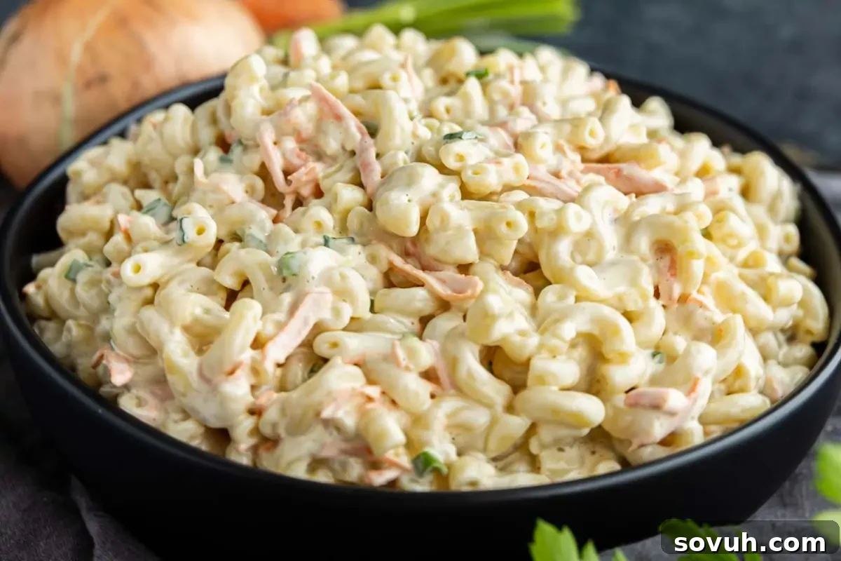A black bowl filled with creamy Hawaiian macaroni salad featuring shredded carrots and chopped herbs, placed on a dark surface with an onion and greens in the background—an easy potluck side dish that’s sure to please any crowd.