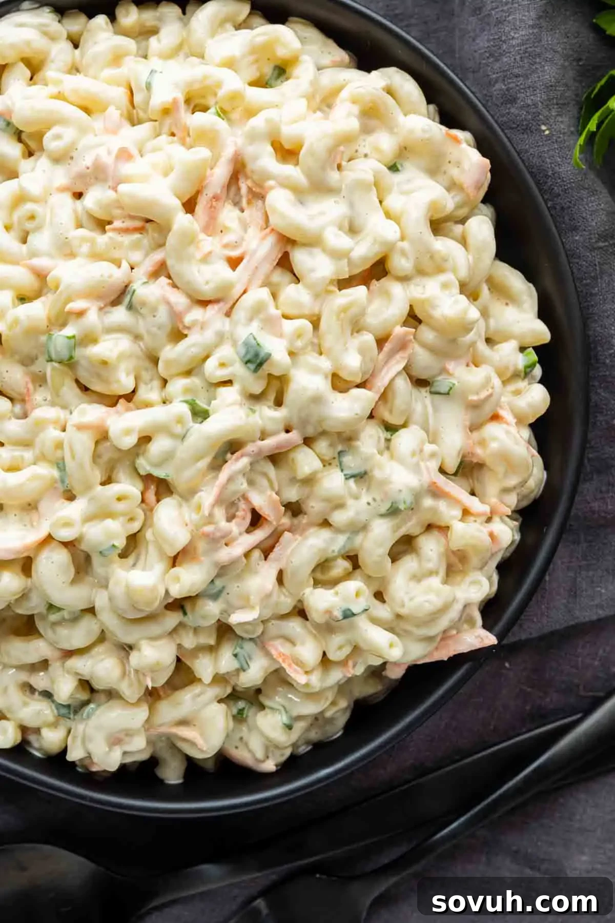 A black bowl filled with creamy Hawaiian macaroni salad—elbow pasta, shredded carrots, and chopped green onions—set on a dark surface. This easy potluck side dish is perfect for any gathering and brings the taste of a classic Hawaiian plate lunch to your table.