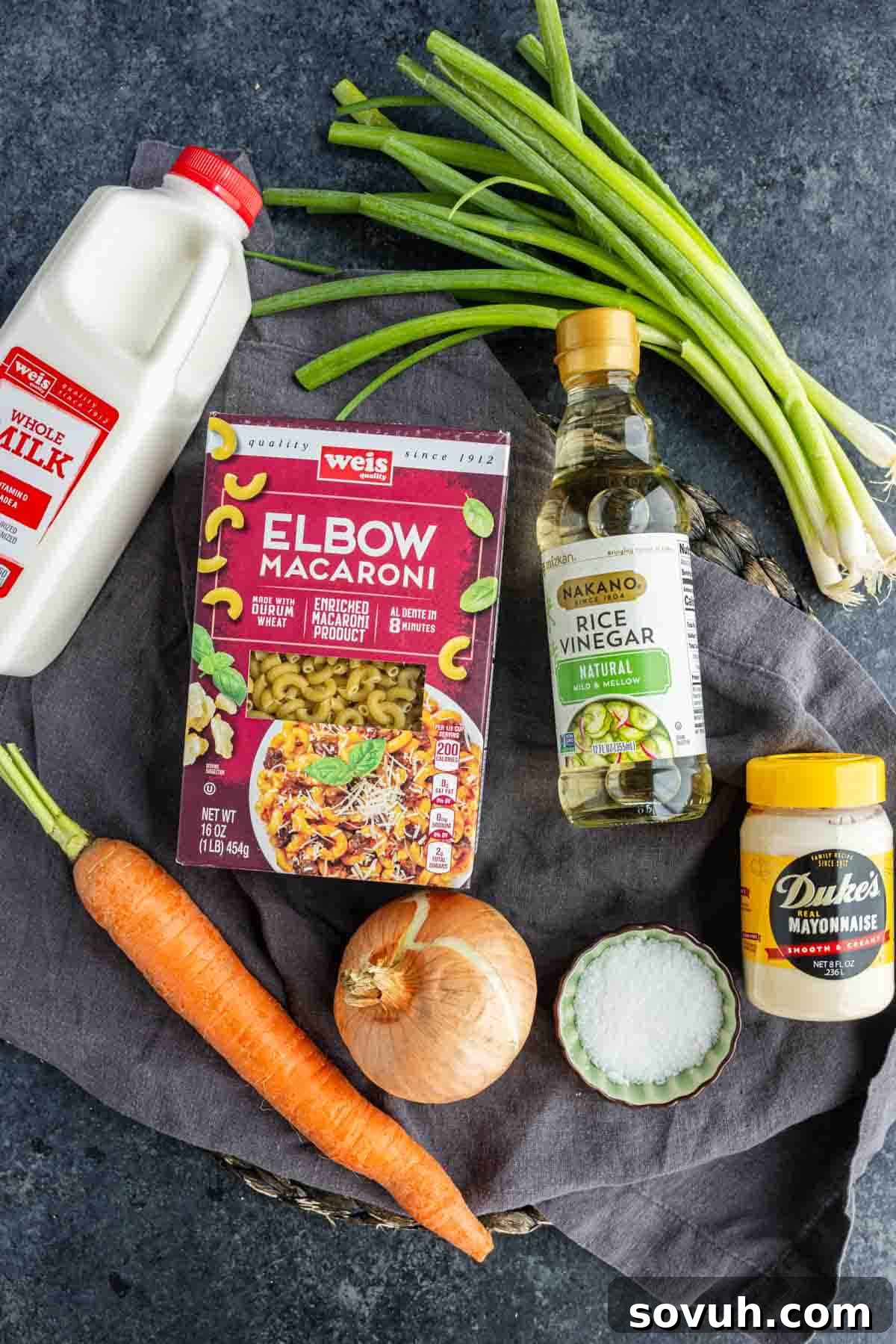A carton of whole milk, elbow macaroni, rice vinegar, green onions, a carrot, a yellow onion, a small bowl of salt, and a jar of mayonnaise are arranged on a dark surface—everything you need to create an easy potluck side dish inspired by classic Hawaiian macaroni salad.