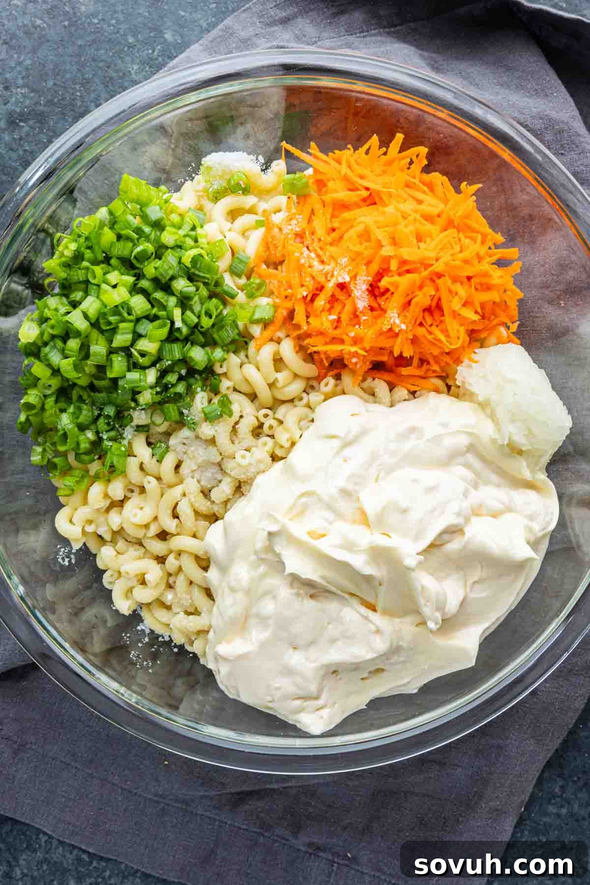 A glass bowl with cooked macaroni, chopped green onions, shredded carrot, mayonnaise, and a scoop of sour cream sits on a dark cloth—a classic Hawaiian macaroni salad perfect as an easy potluck side dish or for serving alongside your favorite Hawaiian plate lunch.