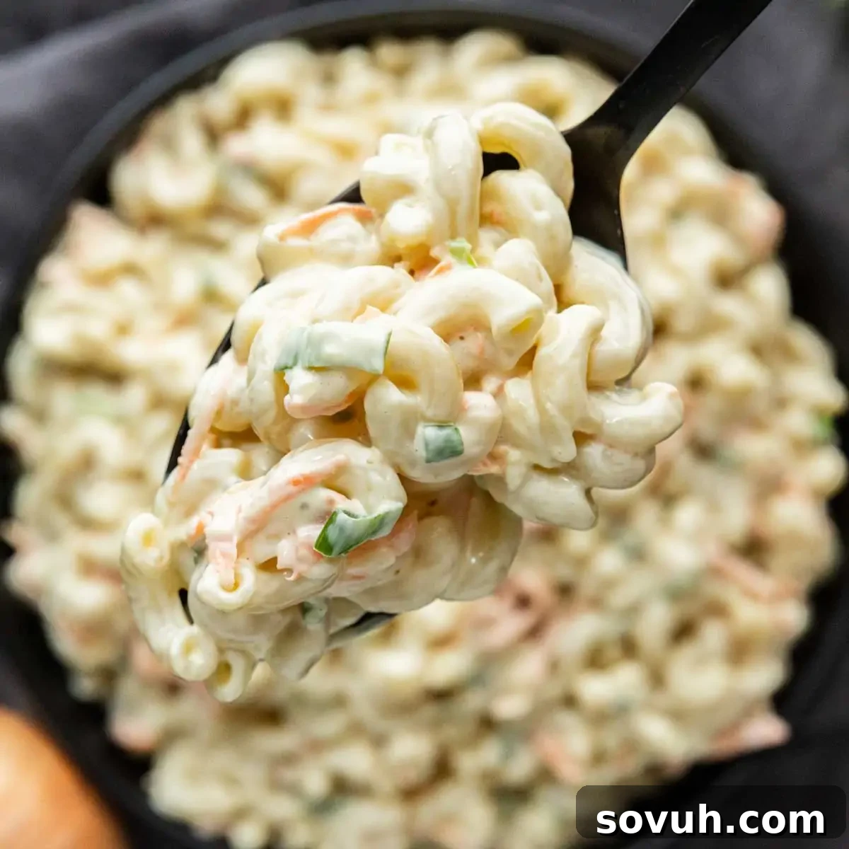 A close-up of a spoonful of creamy macaroni salad with green onions, held above a bowl filled with more macaroni salad—an easy potluck side dish that's always popular at any gathering and a classic addition to a Hawaiian plate lunch.