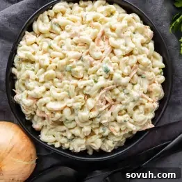 A black bowl filled with Hawaiian macaroni salad—elbow pasta, shredded carrots, green onions, and creamy dressing—sits on a dark cloth with a yellow onion and utensils nearby, making it an easy potluck side dish perfect for any Hawaiian plate lunch.