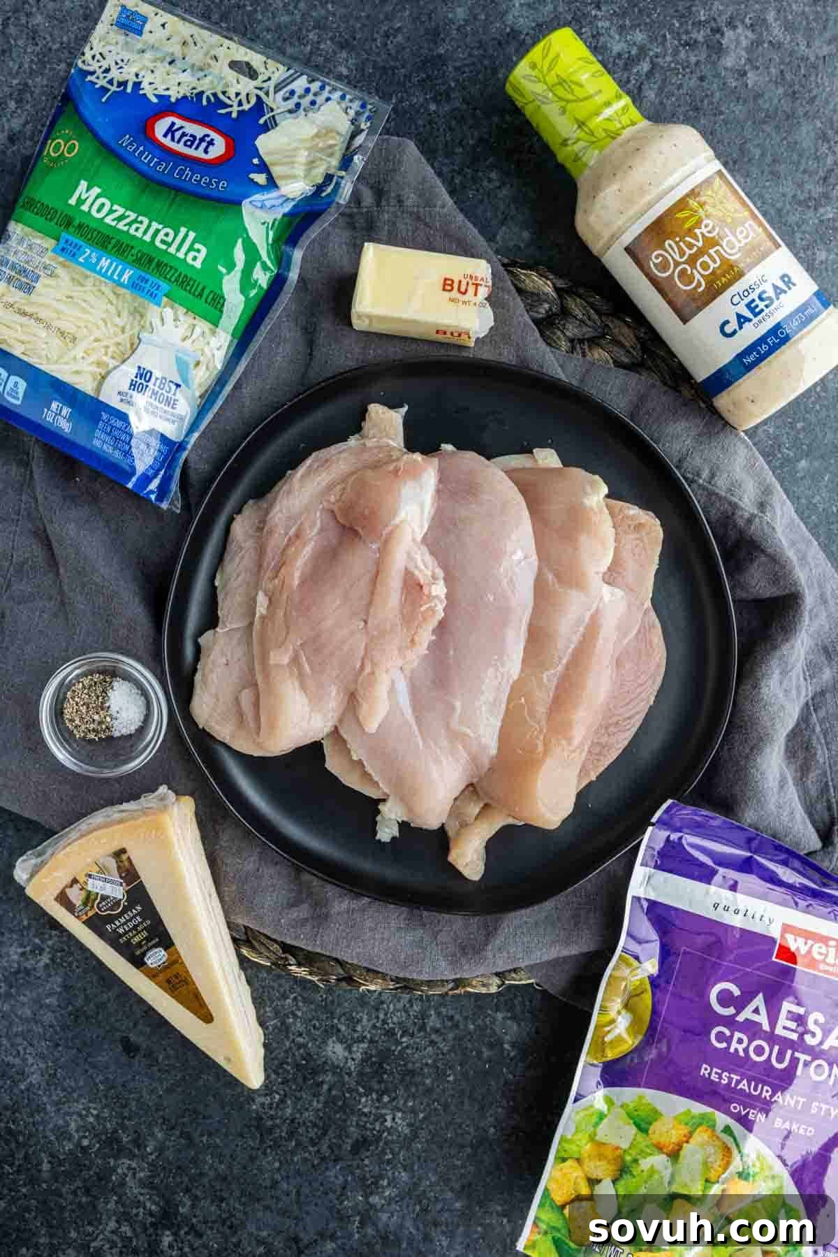 A plate of raw chicken breasts, perfect for baked Caesar chicken or any easy weeknight chicken recipe, is surrounded by mozzarella cheese, Caesar dressing, croutons, Parmesan cheese, butter, and black pepper on a dark surface.