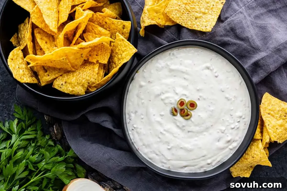 A bowl of tortilla chips next to a bowl of creamy Hamlin's White dip garnished with sliced green olives, placed on a dark cloth with parsley nearby.