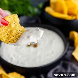 A hand holding a tortilla chip dipped in creamy white sauce, with a bowl of dip and more chips in the background.