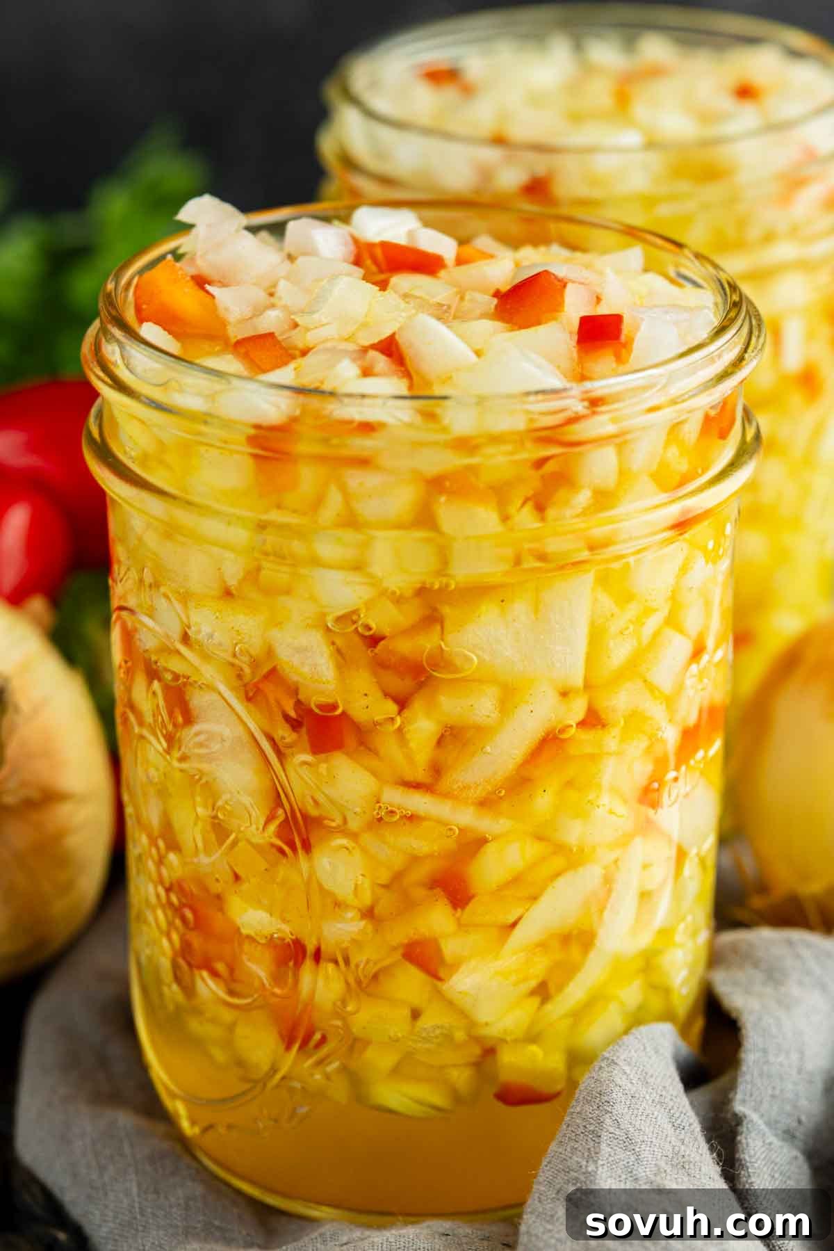 A close-up of two glass jars filled with diced onions and peppers in a yellowish liquid, with vegetables visible in the background.