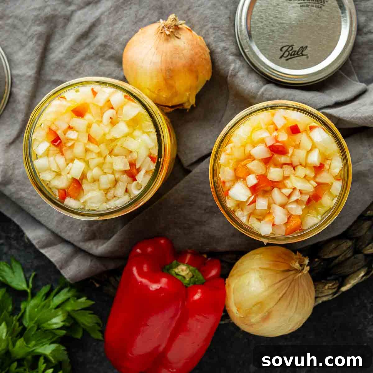 Two jars filled with chopped onions and red peppers on a cloth, surrounded by whole onions, a red bell pepper, parsley, and jar lids.