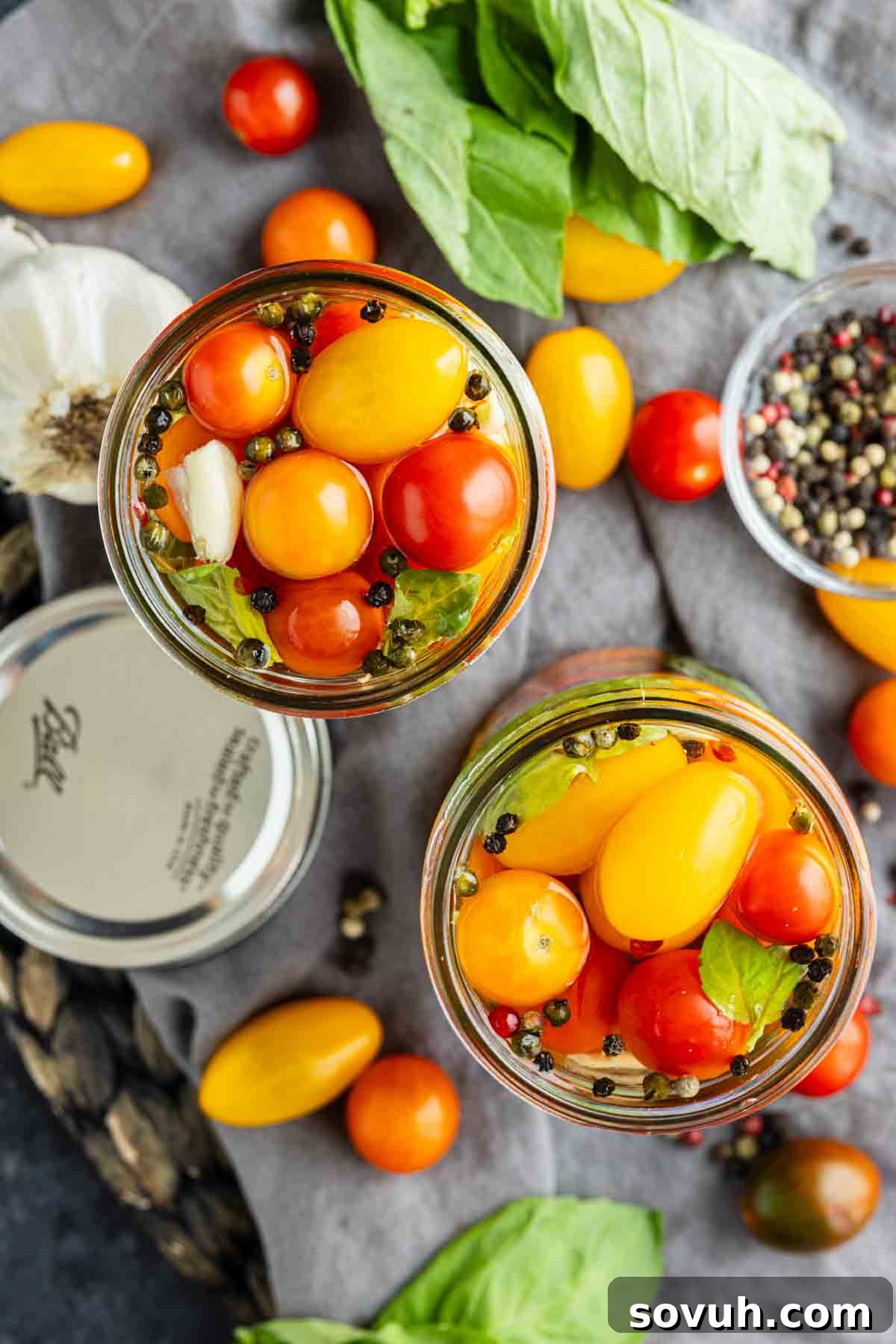 Two glass jars filled with vibrant red and yellow pickled cherry tomatoes, basil leaves, garlic, and peppercorns, surrounded by fresh tomatoes, basil, pepper, and a jar lid on a gray cloth—a perfect inspiration if you’re curious about how to pickle cherry tomatoes at home.